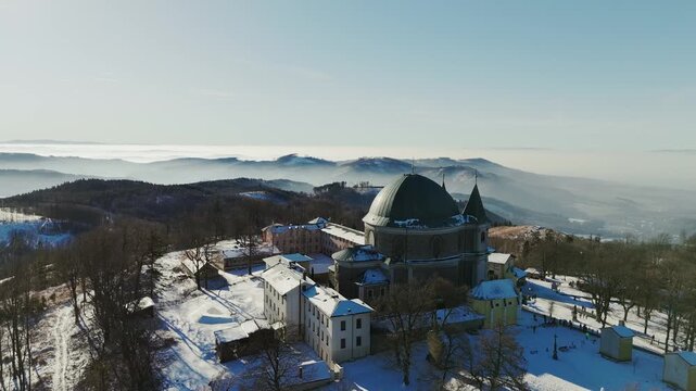 Aerial view of a snow-dusted church and monastery atop a hill, with misty mountains fading into the horizon, Trnava Region, Slovakia.