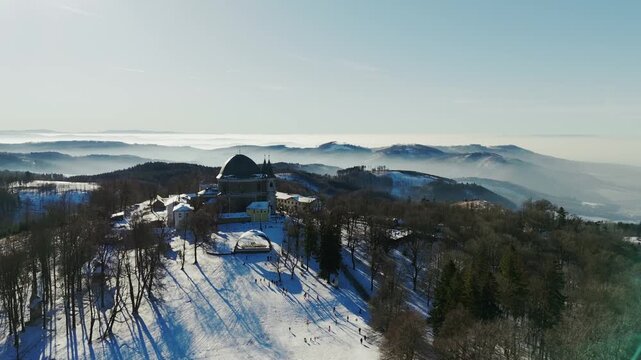 Aerial view of snow-covered hill with a Basilica, skiers enjoying the slopes under a clear sky, Marianka, Trnava Region, Slovakia.