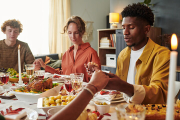 Diverse group of young adults holding hands and praying before meal at dining table, multiethnic friends gathering for holiday celebration with Thanksgiving food and candles