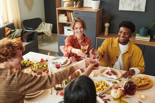 Group of young adults and one Black man sitting around table raising glasses in toast, sharing meal together, smiling and interacting, diverse friends celebrating Thanksgiving occasion indoors