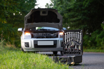 Tool kit on the road in front of a car with an open hood. Open suitcase with keys and tools placed on the asphalt in front of a gray car with the hood open due to a breakdown, technical assistance