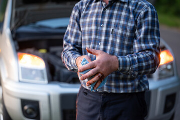 Man in a plaid shirt wiping dirty hands with a rag near an open car hood. A man wipes his hands dirty from engine oil with a rag against the background of a broken car, completion of repairs