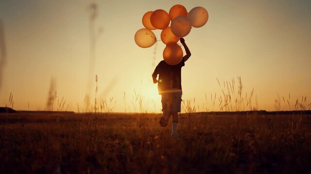 Happy child running over field with balloons in hand, back view, childhood. Beautiful sunset in nature, carefree little boy running to horizon, rear view, cinematic slow motion shot, silhouette