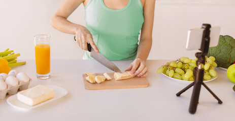 Young blogger preparing fruit salad, cutting banana and recording new video recipe for her food blog on smartphone, standing in kitchen, crop