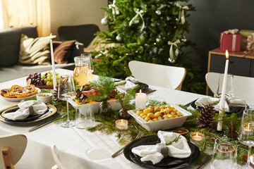 Christmas holiday table setting featuring plates, glasses, candles, pine branches, assorted fruits and dishes arranged on tablecloth with decorated Christmas tree in background