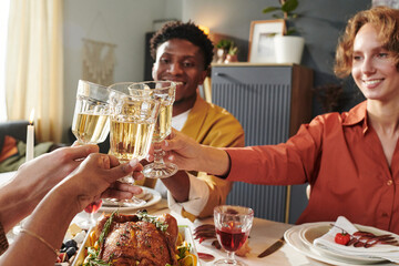 Diverse group of young adults clinking glasses around dining table, celebrating together, smiling and enjoying meal, visible roasted poultry and wine, Thanksgiving gathering atmosphere