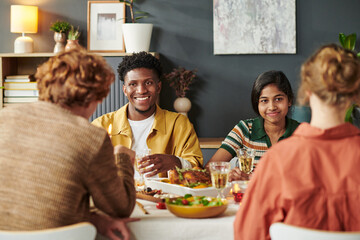 Young Black man and young South Asian woman sitting at dining table with two Caucasian adults, smiling and talking while sharing meal, diverse group enjoying food together indoors