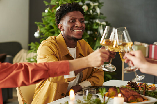 Young adult Black man smiling while clinking wine glasses with friends during Christmas meal, sitting at decorated table with Christmas tree in background, celebrating holiday together - Powered by Adobe