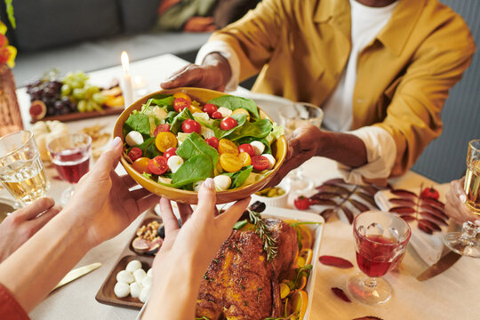 Multiethnic group of young adults sharing fresh salad at Thanksgiving dinner table, hands reaching for bowl, visible roast chicken, glasses of red beverage, assorted fruits in background - Powered by Adobe