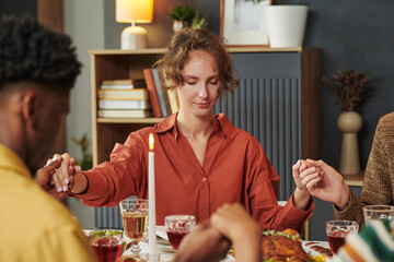 Caucasian young adult woman sitting at table holding hands with diverse group of people, closing eyes in prayer before meal, surrounded by food and drinks during gathering