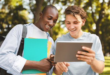 Student guy showing his project on digital tablet to friend, walking outdoors in college campus,...