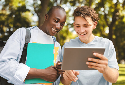 Student guy showing his project on digital tablet to friend, walking outdoors in college campus, looking at device and smiling. University lifestyle concept