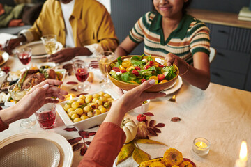 Young Asian woman smiling while passing bowl of fresh salad to another during Thanksgiving meal with friends, multiethnic group enjoying shared dinner at table with various dishes
