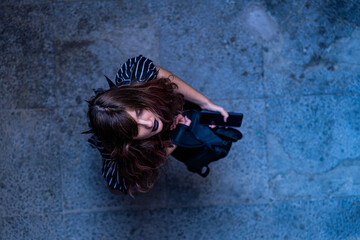 Top view of a young woman in dark clothes holding a phone and backpack, standing on a textured blue pavement, eyes closed, evoking nostalgia and melancholy.
