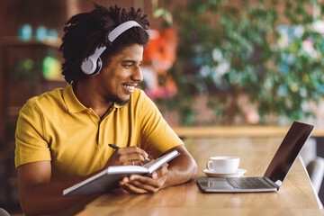 Smiling young black curly man in wireless headphones makes notes in notebook and looks at laptop in cafe interior. Work remotely for digital nomad, modern education and learn language, online lesson