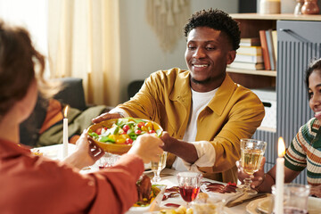Young adult Black man smiling while handing bowl of salad to Caucasian woman during group meal, sitting at dining table with food and drinks, engaging in friendly conversation