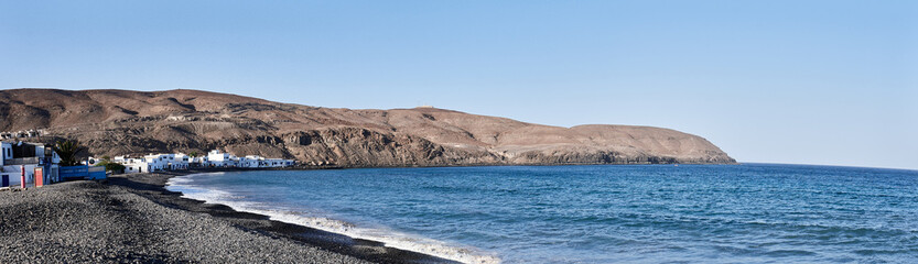 Wide panoramic shot of the rocky coast of Pozo Negro in Fuerteventura, Canary Islands. Blue sky, volcanic landscape and traditional white houses by the sea. Peaceful and natural scenery.