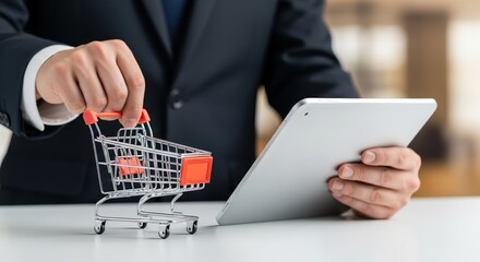 Compras online. A m&atilde;o de um empres&aacute;rio de terno segurando um pequeno carrinho de compras em uma m&atilde;o e um tablet na outra, conceito de com&eacute;rcio eletr&ocirc;nico
