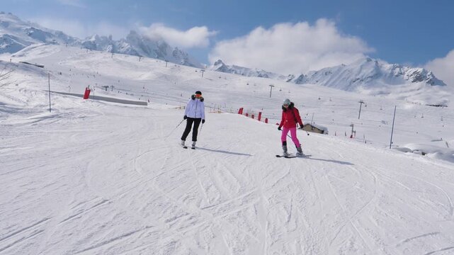 Two female friends in bright ski gear skiing downhill on prepared slope in mountain resort during sunny day. Beautiful alpine scenery with snow covered peaks and blue sky in background.