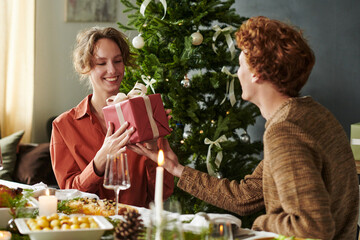 Caucasian young woman smiling while receiving wrapped gift box from Caucasian young man, sitting in front of decorated Christmas tree during Christmas holiday meal at dining table