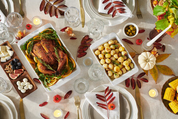 Overhead view showing Thanksgiving table setting with roasted whole chicken, roasted potatoes, corn on cob, cheese platter, nuts, olives, candles, autumn leaves, plates, glassware