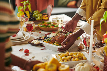 Diverse group of adults gathering around Thanksgiving table, Black man serving roasted turkey while others reaching for food, sharing meal with various dishes and drinks visible