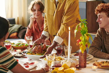 Young Black man serving roast turkey to diverse group of young adults sitting around dining table sharing Thanksgiving meal, enjoying food and conversation together
