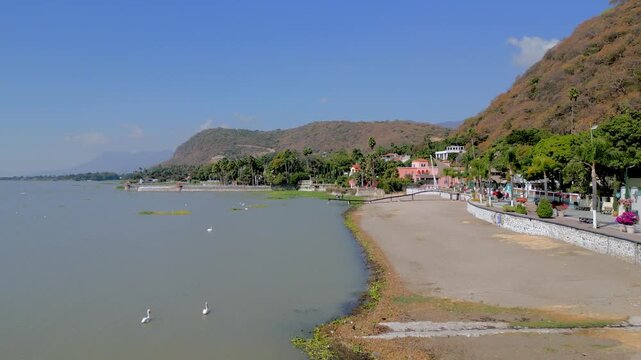 Aerial drone footage gliding beside the Chapala boardwalk with lakeside homes and hills in Jalisco, Mexico
