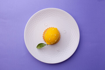 Overhead shot of a hyper-realistic lemon-shaped dessert: textured surface, dew-like droplets, and green leaf on a ridged white plate against purple.
