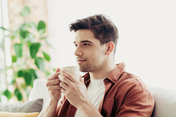 Fototapeta premium Young man enjoying morning coffee at home, sitting comfortably on a sofa in a bright modern interior