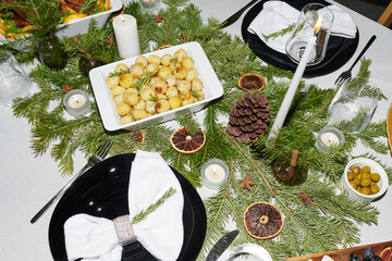 Christmas table setting featuring evergreen branches, pine cone, dried citrus slices, candles, black plates with napkins, bowl of roasted potatoes and dish of green olives