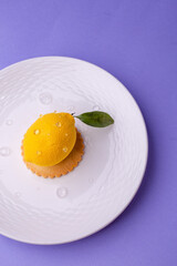 Overhead shot of a hyper-realistic lemon-shaped dessert: textured surface, dew-like droplets, and green leaf on a ridged white plate.