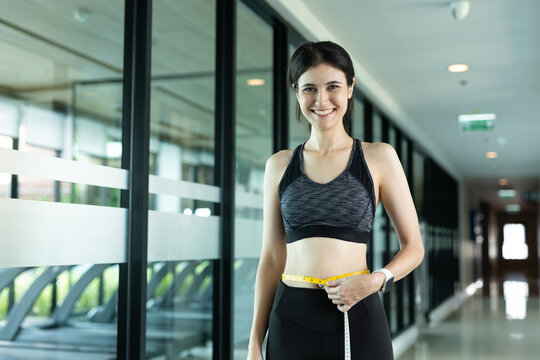Smiling women in sportswear measuring her waist with tape measure in gym hallway, symbolizing weight loss progress, fitness goals, body confidence and healthy lifestyle.