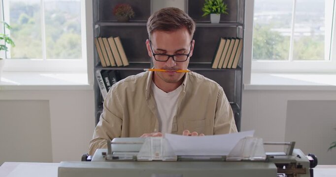 Busy writer typing on vintage typewriter in office by windows, holding pencil in mouth. Man wearing glasses for focus and creativity of retro vibes, professional authors routine working on old machine - Powered by Adobe