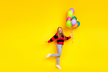 Cheerful young girl holding colorful balloons in front of a bright yellow background, expressing...