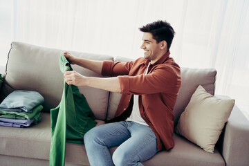 Young man happily folding clothes while sitting on a cozy couch in a bright interior, reflecting...