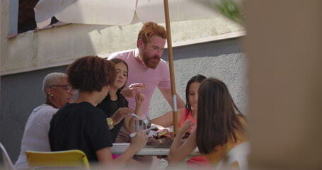 Friends dining together outdoors under an umbrella, sharing laughter, food, and conversation,...