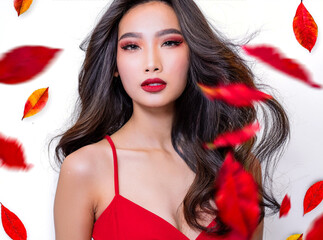 A woman with dramatic makeup and a red dress is framed against a white background, with red leaves swirling around her. She is looking directly at the camera.