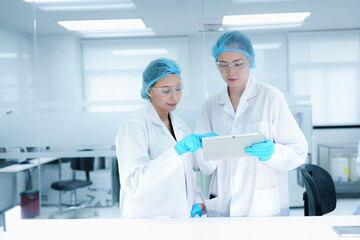 A senior scientist mentors a junior colleague in a modern lab, using a tablet to guide her through...