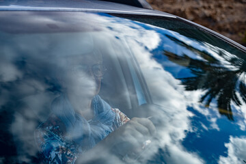 Concentrated senior woman driving a car, seen through the windshield. Confident elderly lady driver wearing glasses sits behind the wheel. Concept of independence, freedom, and transportation.