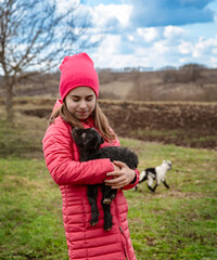 Smiling girl in red jacket and beanie holding black baby goat on farm. Rural lifestyle, childhood, animal care countryside setting