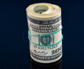 Rolled stack of hundred dollar bills secured with a rubber band, resting on a reflective surface,...