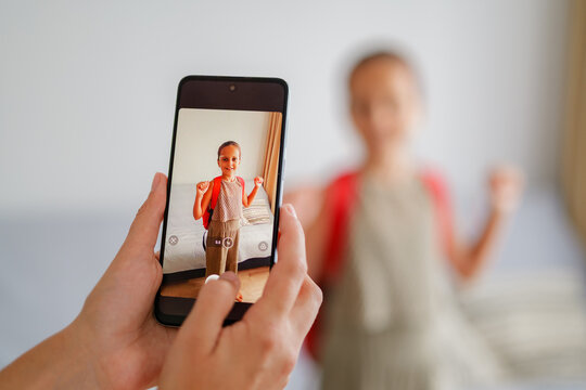 
A close-up of a smartphone screen shows a mother taking a picture of her smiling daughter posing with a red backpack on her first day of school.
