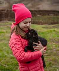 Smiling girl in red jacket and beanie holding black baby goat on farm. Rural lifestyle, childhood, animal care countryside setting