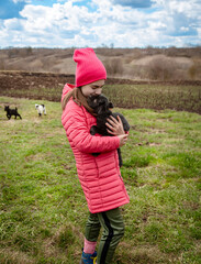 Smiling girl in red jacket and beanie holding black baby goat on farm. Rural lifestyle, childhood, animal care countryside setting