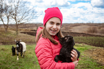 Smiling girl in red jacket and beanie holding black baby goat on farm. Rural lifestyle, childhood, animal care countryside setting