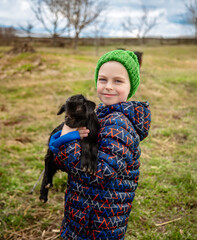 Joyful boy holding black baby goat on farm. Rural lifestyle, childhood, and animal care in countryside setting. Happy childhood