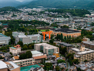 Obraz premium Aerial view of Kyoto, Japan, featuring a large orange torii gate, traditional temple complex, modern buildings, and forested hills in the background.