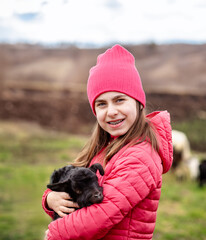 Cute girl in red jacket and beanie holding black baby goat on farm. Rural lifestyle, childhood, animal care countryside setting