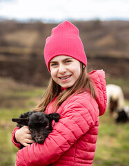 Cute girl in red jacket and beanie holding black baby goat on farm. Rural lifestyle, childhood, animal care countryside setting